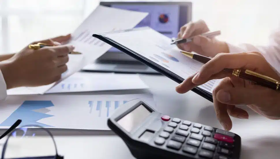 Close-up of hands working with financial charts, a calculator, and documents at a desk, representing accounting or financial analysis work.