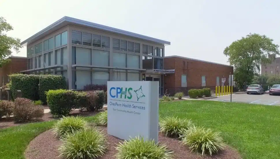 Exterior view of ChesPenn Health Services building with modern glass and brick architecture, landscaped greenery, and a sign reading "ChesPenn Health Services – Your Community Health Center.