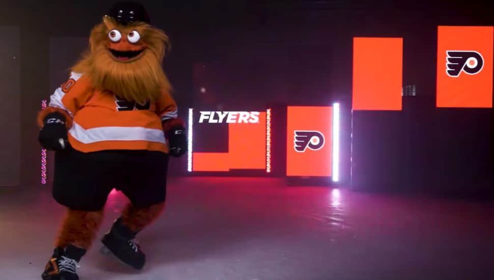 Philadelphia Flyers mascot Gritty skating on an ice rink with bright orange Flyers logo screens in the background.