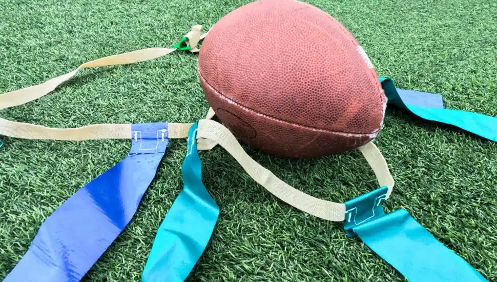 A close-up of a football and flag belts lying on a turf field, representing flag football equipment used in non-contact play.
