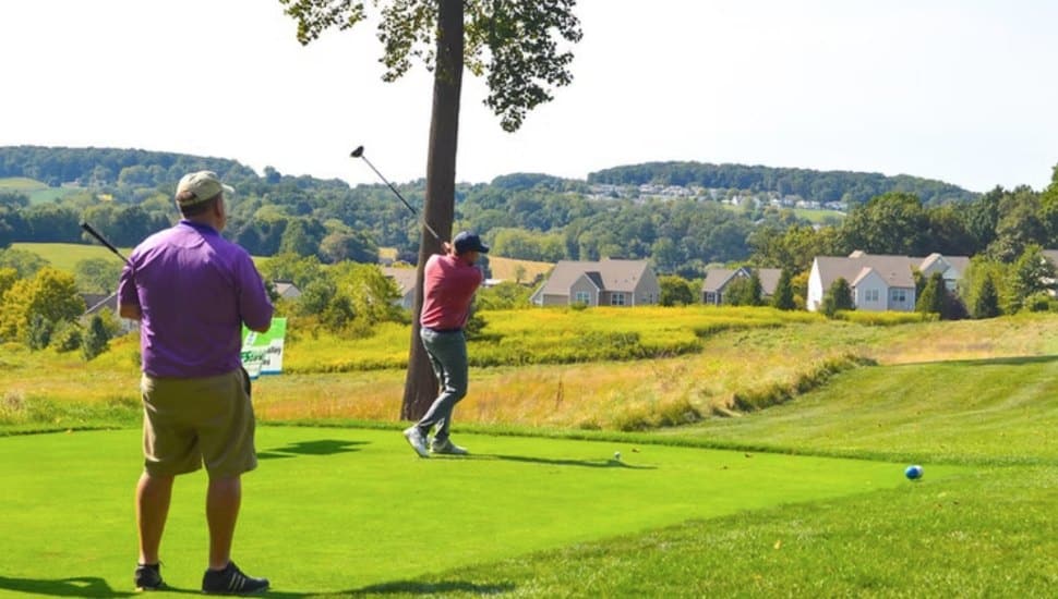 Two golfers on a sunny green at French Creek Golf Club, with one mid-swing and the other watching. Rolling hills and suburban homes are visible in the background.