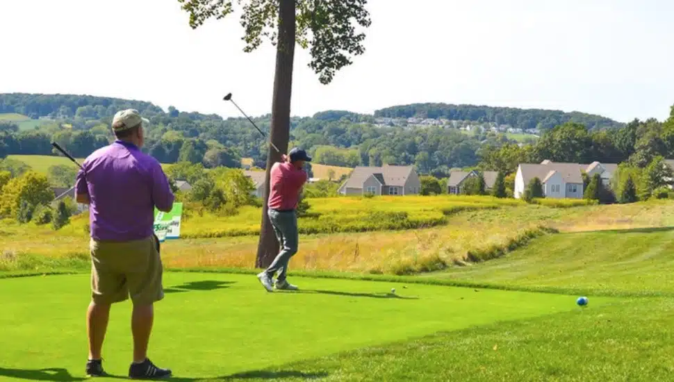 Two golfers on a sunny green at French Creek Golf Club, with one mid-swing and the other watching. Rolling hills and suburban homes are visible in the background.