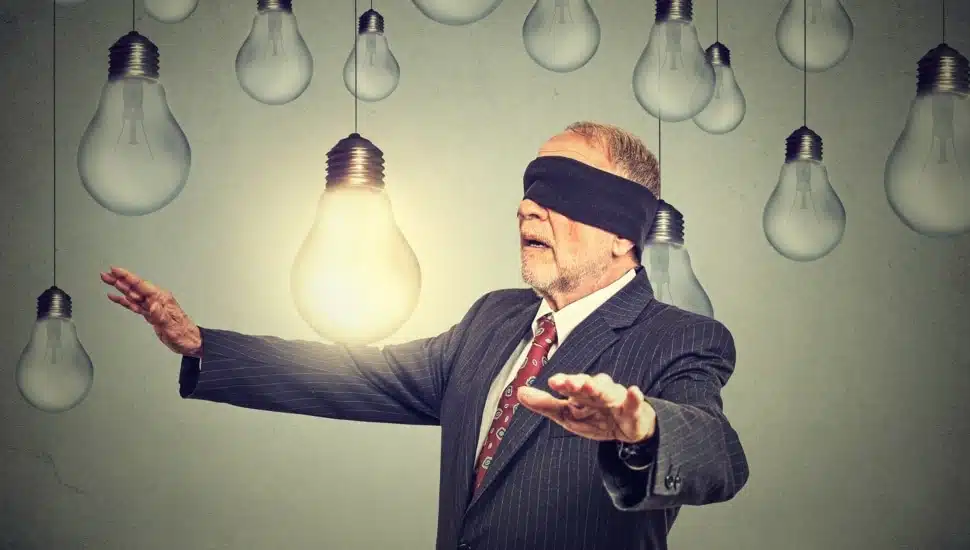 Blindfolded man walking through light bulbs searching for bright idea.