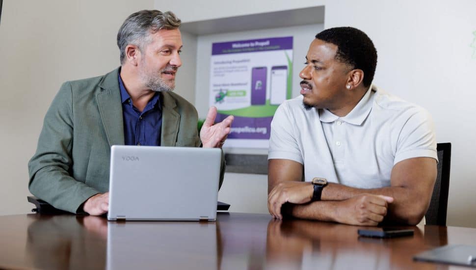 Two men sitting at a conference table having a focused discussion, with a laptop open in front of them and a Propell Credit Union promotional poster visible in the background.