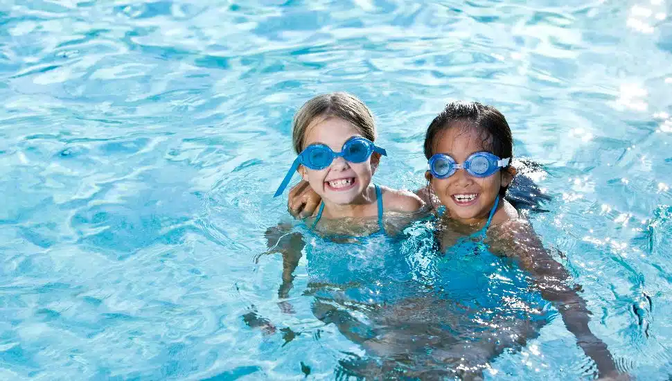 Two smiling young girls wearing swim goggles hug each other while standing in a clear blue pool, enjoying a swim on a sunny day.