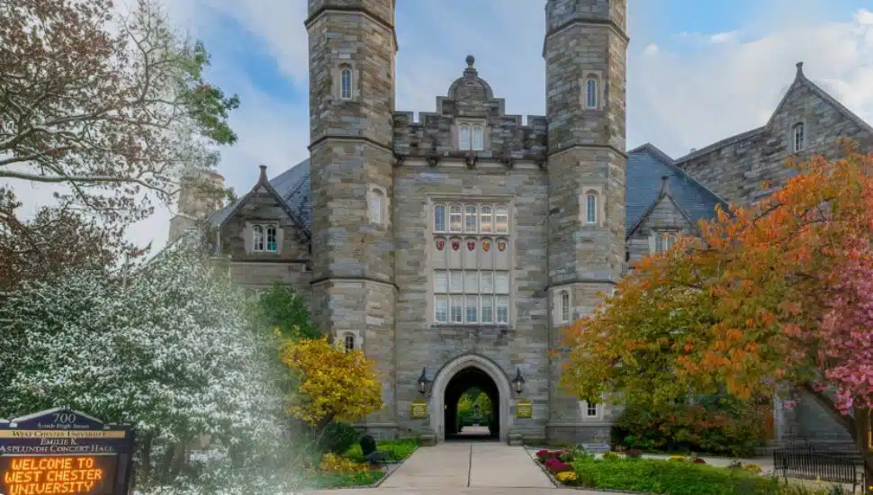 Stone archway entrance of West Chester University’s Philips Memorial Building, framed by trees in bloom and fall foliage, with a sign welcoming visitors to campus.