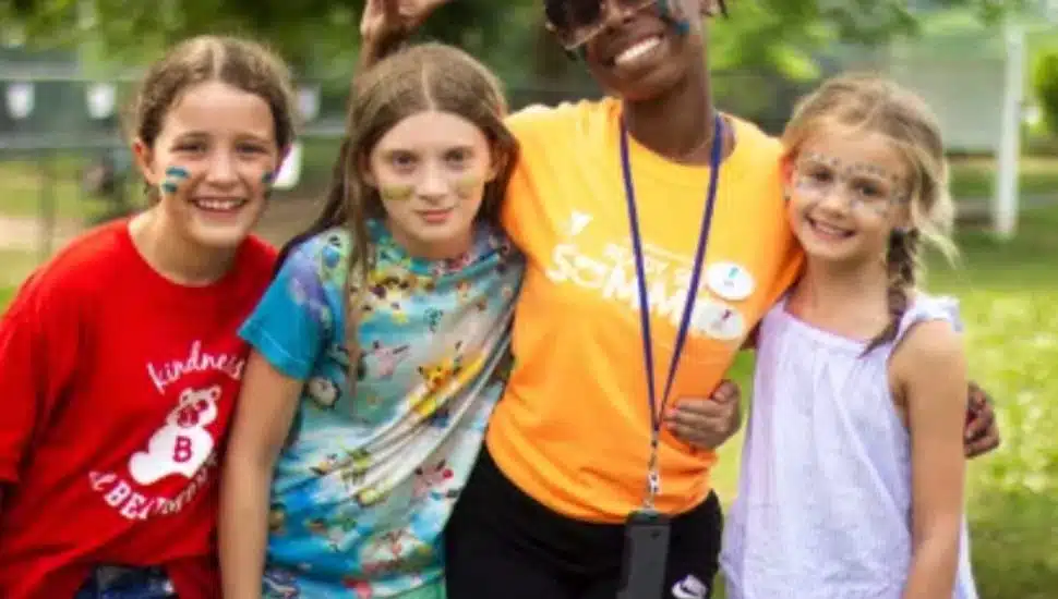 A YMCA summer camp counselor smiles with three young campers, all with face paint and arms around each other, enjoying outdoor activities in a cheerful and inclusive setting.