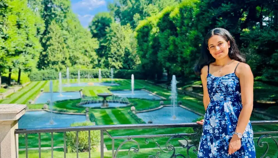 Young woman in a blue floral dress standing by a decorative railing with an elegant garden and fountains in the background.