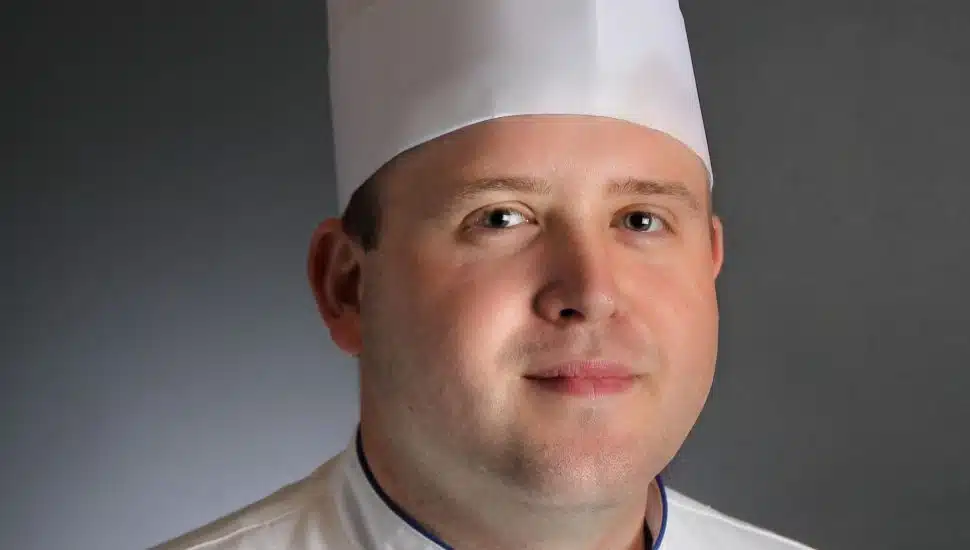 Portrait of Chef Jonathan Fike wearing a traditional white chef's uniform and tall pleated chef hat. His coat is embroidered with his name and professional titles: “Jonathan Fike, CEC, CCA."