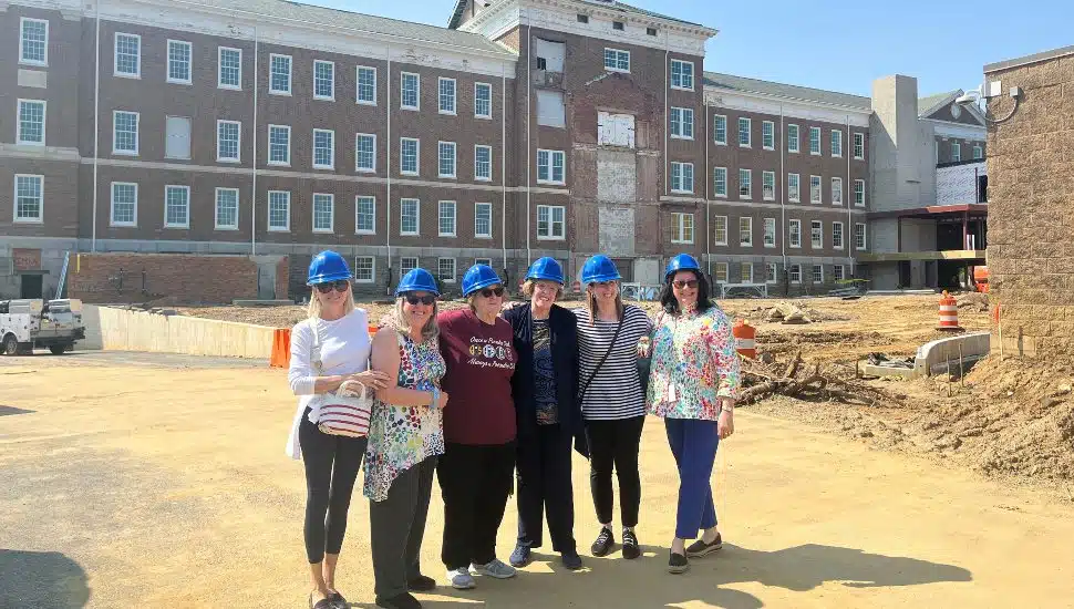 Group of Archbishop Prendergast High School alumnae wearing blue hard hats standing in front of the historic school building under renovation, soon to become the DCCC Southeast Center.