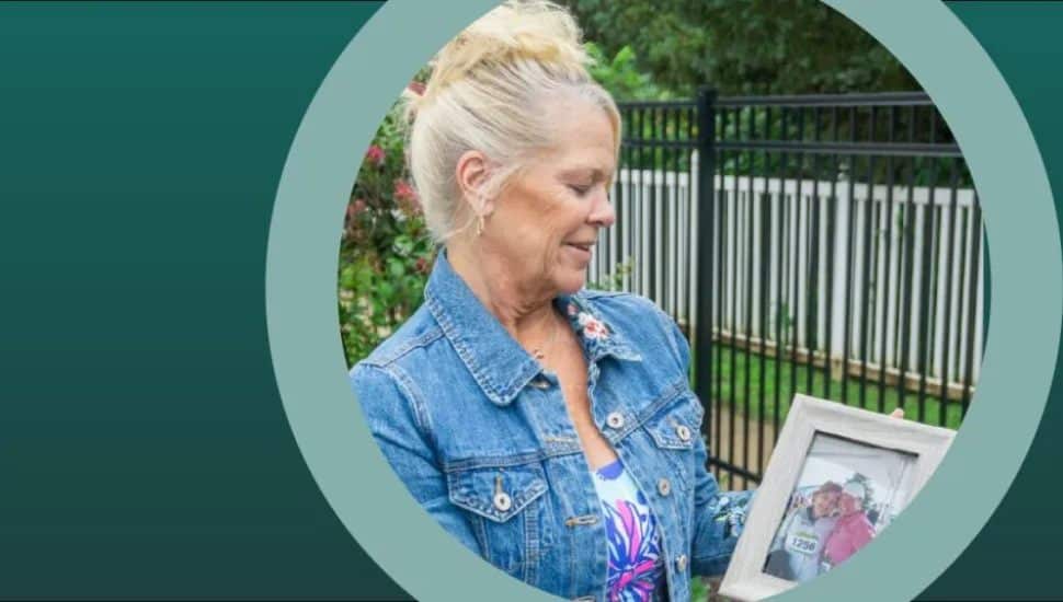 Smiling woman in a denim jacket holds a framed photograph of two people at an event, standing outdoors near a black fence and greenery.