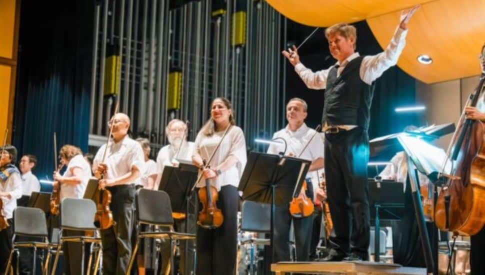 Brandywine Valley Symphony musicians standing with their instruments on stage as Music Director Michael Hall raises his arms to acknowledge the audience, with a large pipe organ in the background.