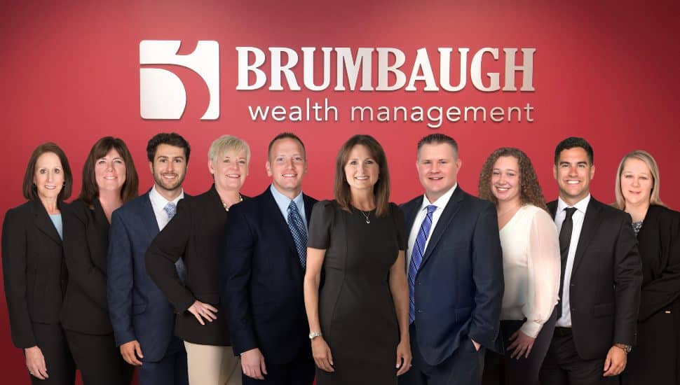 Group photo of the Brumbaugh Wealth Management team standing in front of a red wall with the company logo, dressed in professional business attire.