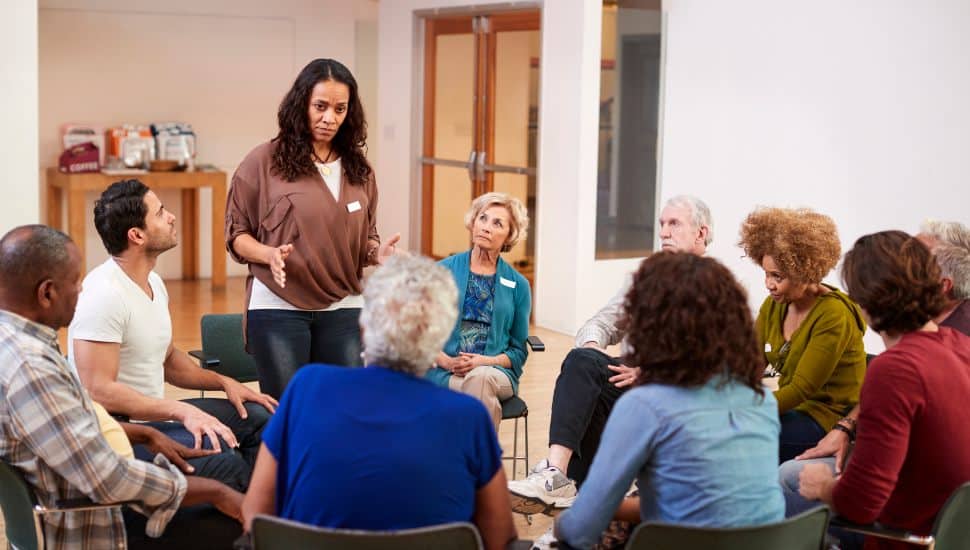 A diverse group of adults sits in a circle during a community health and wellness support group, with one woman speaking while others listen attentively in a bright meeting room.