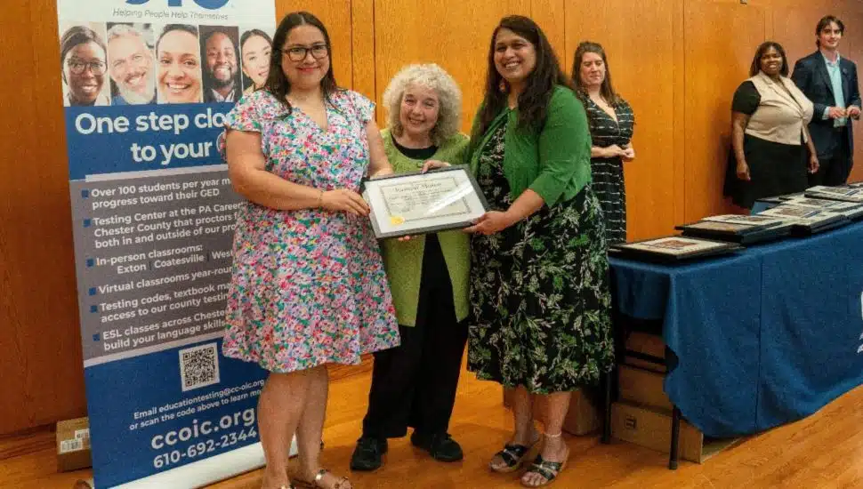 Three women smiling and holding a graduation certificate at a Chester County OIC ceremony, with staff and awards table in the background.