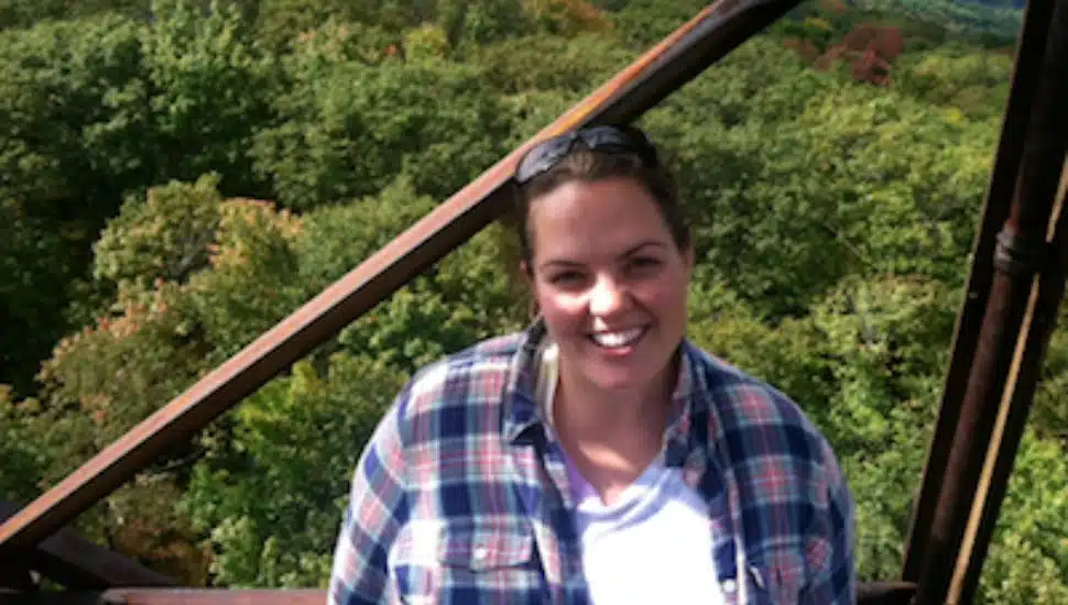 Smiling woman in plaid shirt standing on a metal structure with green trees in the background.