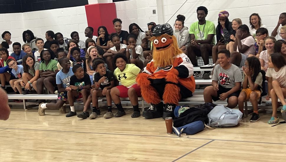 Philadelphia Flyers mascot Gritty sits with a large group of excited YMCA summer campers in a gymnasium, surrounded by smiling children and staff during a youth engagement event at the Coatesville YMCA.