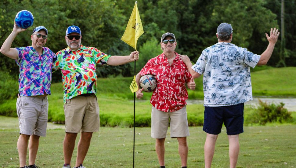 Four guys wearing Hawaiian shirts on golf course holding soccer ball.