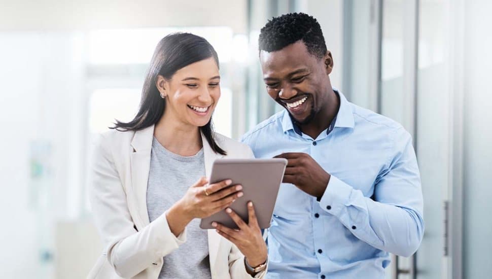 Two professionals smiling while reviewing information on a tablet in a modern office hallway.
