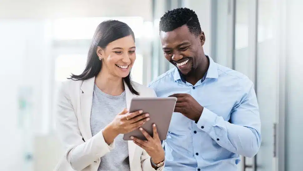 Two professionals smiling while reviewing information on a tablet in a modern office hallway.