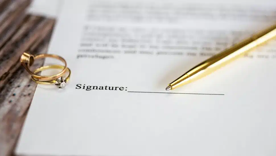 Close-up of a prenuptial agreement with signature line, gold wedding rings, and pen on wooden table.