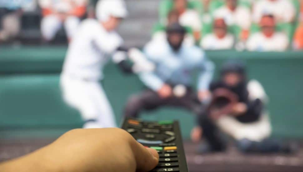 Man clicks remote and watches baseball on blurred tv in background.