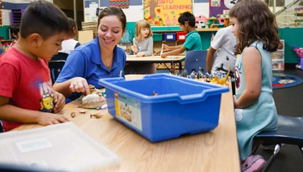 A YMCA staff member smiles while engaging with a young boy and other children at a classroom table filled with toys and learning materials. The inclusive environment reflects adaptive programming for children with developmental delays.