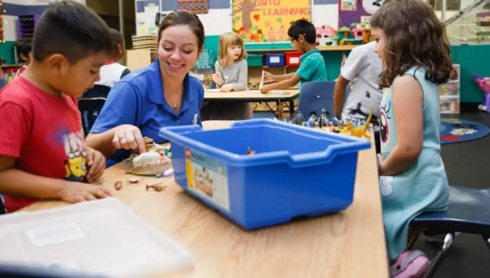 A YMCA staff member smiles while engaging with a young boy and other children at a classroom table filled with toys and learning materials. The inclusive environment reflects adaptive programming for children with developmental delays.
