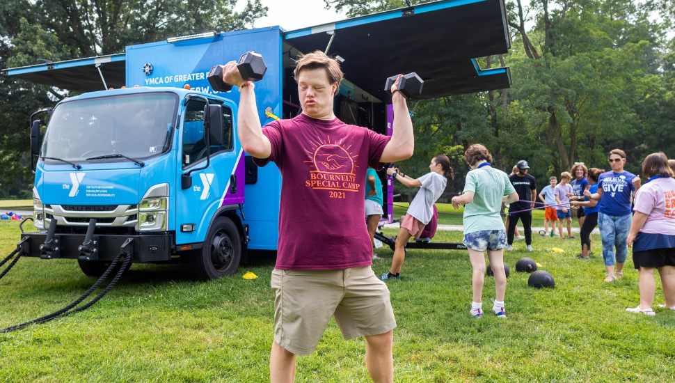 A young man stands on grass lifting dumbbells with both arms in front of a bright blue YMCA mobile fitness truck. He wears a maroon "Bournelyf Special Camp 2021" shirt and khaki shorts, appearing focused and determined. Behind him, a group of people of various ages and abilities engage in outdoor fitness activities, including resistance training and team exercises, surrounded by trees in a park setting.
