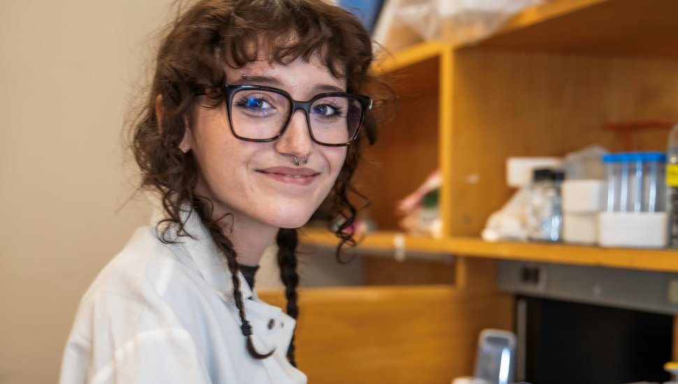 Delaware County Community College student wearing a lab coat and glasses smiles while working in a laboratory with scientific equipment in the background.