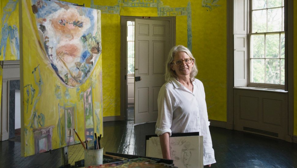 Woman wearing glasses and a white shirt stands in a brightly painted yellow art studio beside an easel with a colorful painting and sketchbooks in hand.