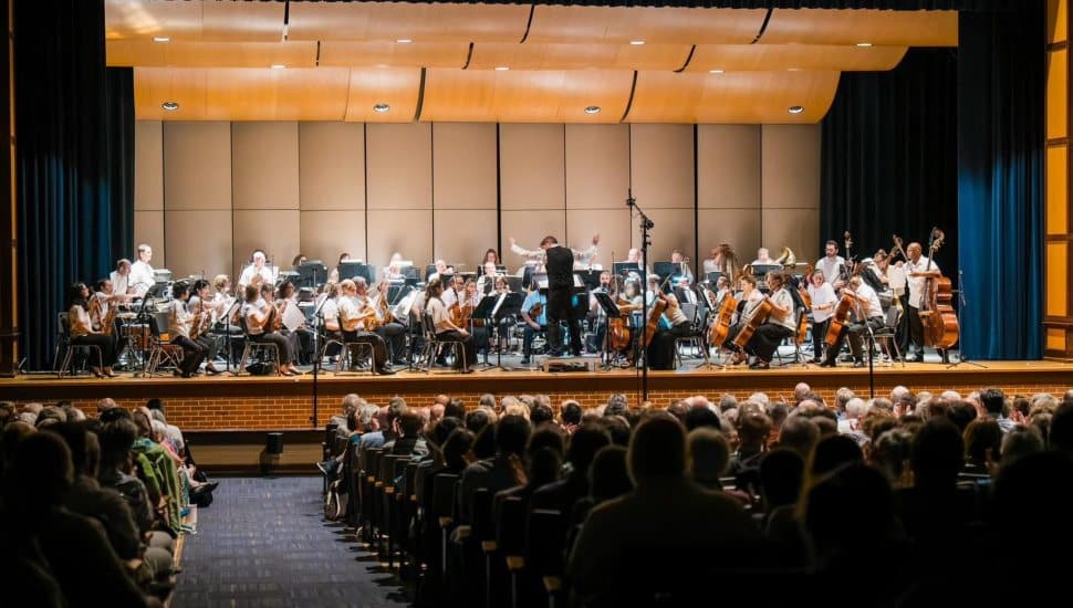 Full orchestra of the Brandywine Valley Symphony performing on stage in front of a large audience, with the conductor leading musicians playing strings, woodwinds, brass, and percussion.