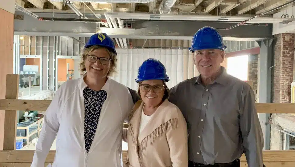 Three people wearing blue hard hats stand inside a construction site. From left to right: Dr. Marian Kozachyn, Vice President at DCCC, Sharon Kelleher, and Andrew Kelleher. They are smiling while visiting the future Southeast Center in Drexel Hill, PA.