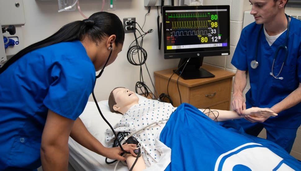 "Two nursing students in blue scrubs practice patient care on a medical manikin, checking vital signs and monitoring a heart rate display on a screen in a clinical simulation lab.