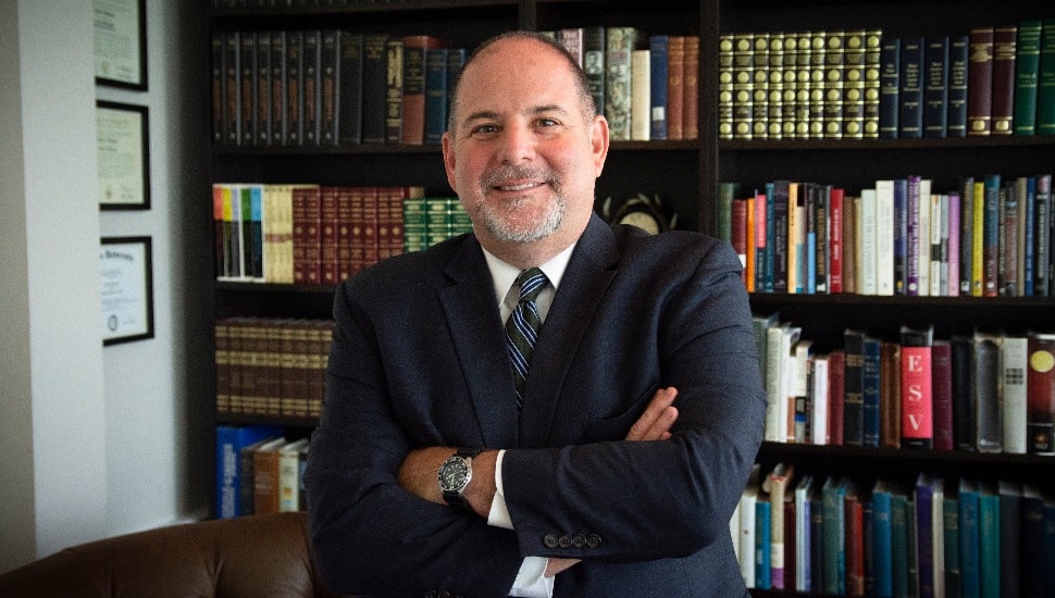 Todd Williams stands in front of a bookshelf in a tie and jacket, smiling with arms crossed.