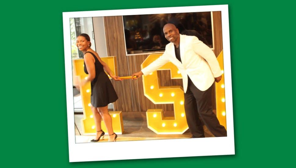 Veda Davida Wooley in a black dress and her father, Dave Wooley, in a white jacket pose playfully while holding a baton in front of large illuminated letters.