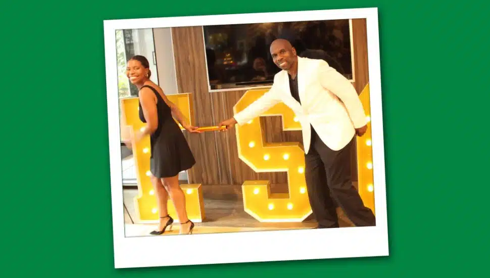 Veda Davida Wooley in a black dress and her father, Dave Wooley, in a white jacket pose playfully while holding a baton in front of large illuminated letters.