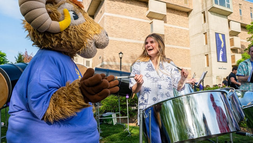 West Chester University mascot Rammy wearing a purple shirt interacts with a smiling student playing steel drums during Concert on the Quad, with Main Hall in the background.