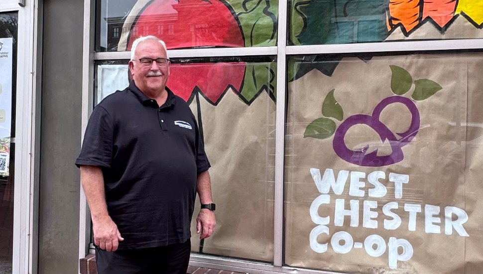 Man in a black Brandywine Valley HVAC polo shirt smiling while standing in front of the West Chester Co-op storefront window decorated with colorful produce artwork.