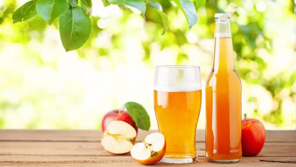 A glass and bottle of hard cider alongside several red apples.