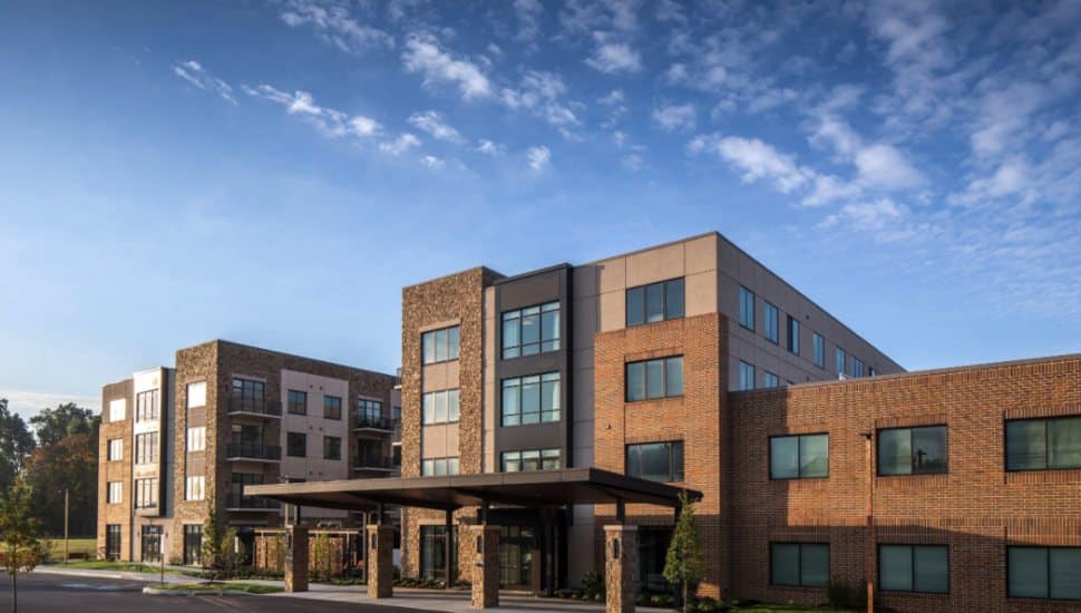 Exterior view of Eagleview Landing Senior Living Community in Exton, Pennsylvania. The modern multi-story building features brick and stone facades, large windows, and a covered entrance, set against a blue sky with scattered clouds.