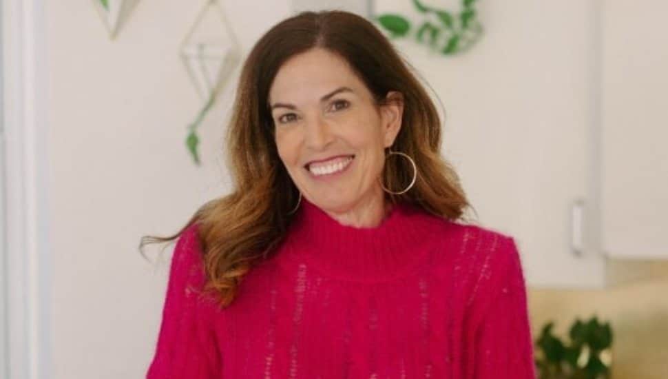 Smiling woman with long brown hair wearing a bright pink sweater and hoop earrings, standing indoors with plants in the background.