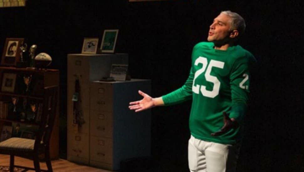 Actor performing in the play “Tommy and Me” at People’s Light, wearing a green football jersey with the number 25, standing on stage near a set decorated with sports memorabilia and family photos.