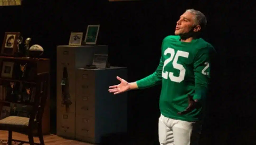 Actor performing in the play “Tommy and Me” at People’s Light, wearing a green football jersey with the number 25, standing on stage near a set decorated with sports memorabilia and family photos.