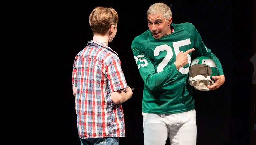 A young actor in a red-and-blue plaid shirt faces an actor dressed as Philadelphia Eagles player Tommy McDonald, wearing a green #25 football uniform and holding a helmet, during a scene from the play "Tommy and Me" at People’s Light.