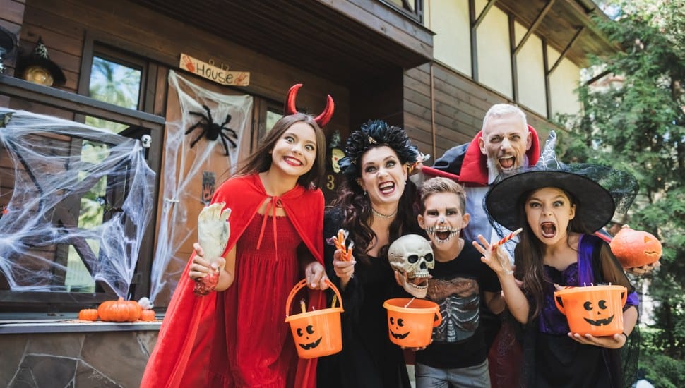 Two adults and several young children in Halloween costumes holding Jack-O-Lantern buckets in front of a decorated door.