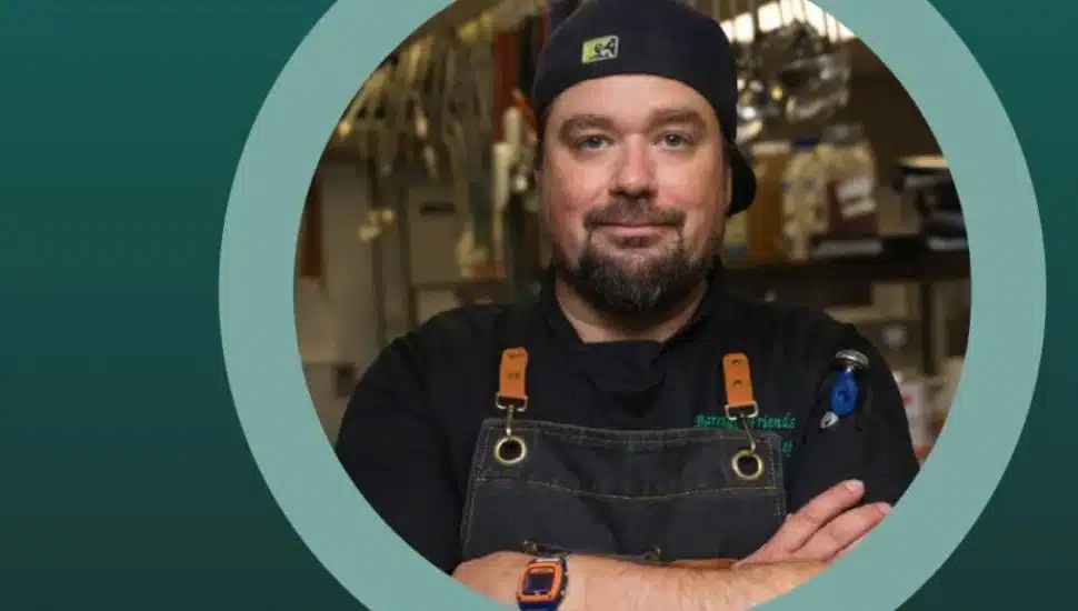 Head Chef Evan Neal of Barclay Friends standing confidently in a commercial kitchen, wearing a dark chef’s uniform and cap.