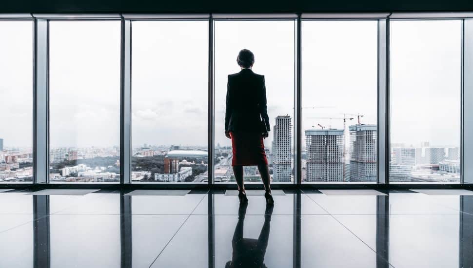 Business woman stands in front of large windows with her back to camera.