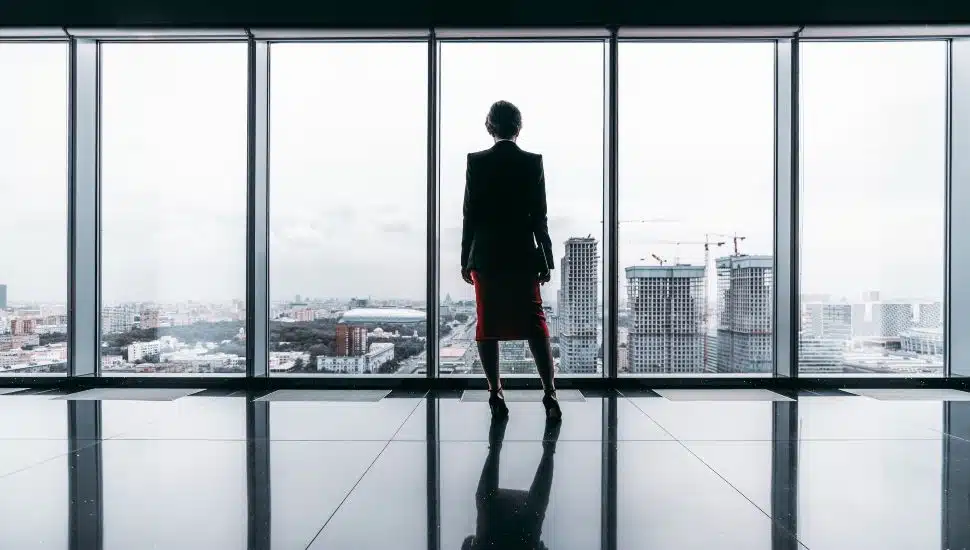 Business woman stands in front of large windows with her back to camera.