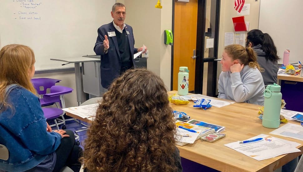 A man stands at the front of a classroom speaking to a group of high school students seated around a table. Papers, worksheets, and snacks are spread out as the students listen attentively during a financial literacy workshop.